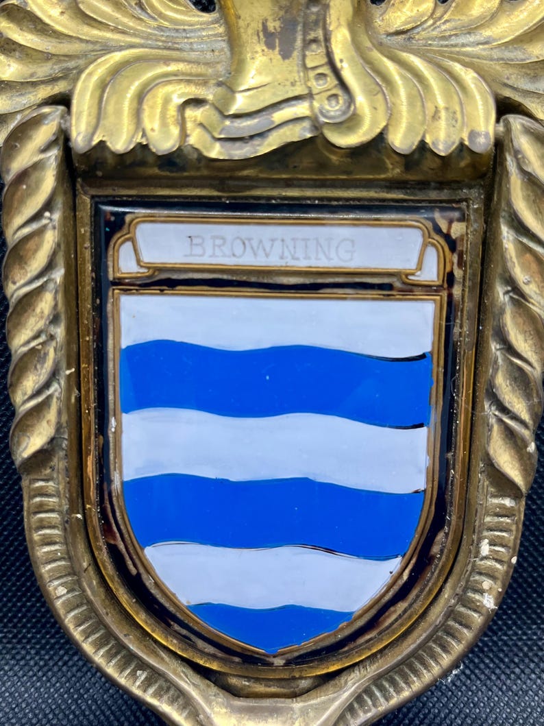 K&ouml;nnte beinhalten: Ein heraldischer Schild aus Messing mit einem zentralen Emaille-Wappen, das abwechselnd blaue und wei&szlig;e, gewellte horizontale Streifen aufweist. Das Wort BROWNING steht auf einem wei&szlig;en Banner oben auf dem Schild, der von einem gedrehten Metallrand umrahmt ist.