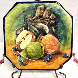 May include: Hand-painted ceramic plate with a vibrant fruit still life. The octagonal plate features a blue border and a colorful arrangement of figs, apples, pears, and blueberries. The plate is displayed on a decorative metal stand.