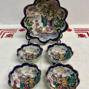 May include: A set of five porcelain serving dishes. The large, scalloped bowl sits on a black metal stand, with four smaller bowls below. Each piece features a hand-painted scene with figures, trees, and mountains, framed by a blue border with gold trim.