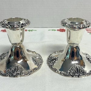May include: A pair of silver-coloured metal candle holders featuring a polished finish, a flared base, and a cylindrical top. The edges of the base and the top rim are decorated with a raised, intricate embossed floral and scroll pattern.