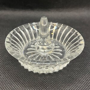 May include: A clear glass ring holder featuring a circular dish base with a radiating sunburst pattern and vertical fluted edges. A central, smooth, conical post rises from the middle of the dish to provide a secure stand for rings.