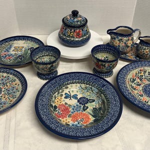May include: A collection of blue and white ceramic tableware with floral patterns. The set includes plates, bowls, cups, a sugar bowl, and pitchers. The pieces have a dark blue rim and are decorated with colorful flowers and butterflies.
