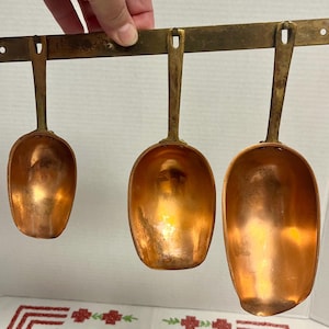 May include: A set of three copper scoops with brass handles hanging from a horizontal brass wall-mounted rack. The scoops vary in size, each featuring a deep, rounded bowl and a long, flat handle attached to the rectangular metal bar.