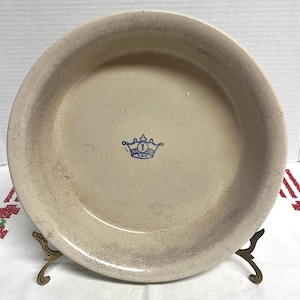 May include: A round, beige ceramic pie dish with a visible crackle glaze finish. The center of the dish features a blue stamped logo of a crown with the number 1 and the text U.S.A. inside. The dish is supported by a decorative metal stand.