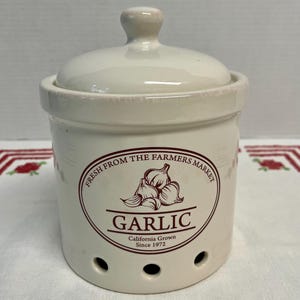 May include: Cream-coloured ceramic garlic storage pot with a lid and handle. The pot features a circular design with the words "FRESH FROM THE FARMERS MARKET" and "GARLIC" in brown. It also states "California Grown Since 1972".