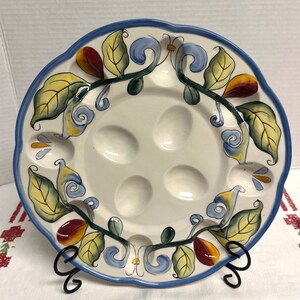 May include: A round ceramic egg serving plate with a scalloped blue rim. The center features four recessed egg-shaped indentations. The border is decorated with hand-painted green and yellow leaves, blue swirls, and small red fruit accents.