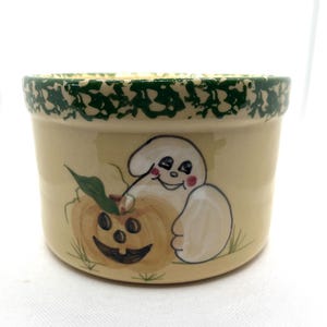 May include: A ceramic Halloween-themed bowl with a cream-colored base and a green speckled rim. The bowl features a hand-painted design of a smiling ghost and a jack-o'-lantern. The bowl is suitable for serving snacks or as a decorative piece.