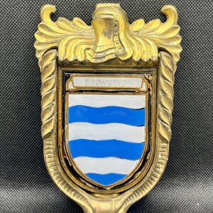 K&ouml;nnte beinhalten: Ein T&uuml;rklopfer aus Messing mit einem schildf&ouml;rmigen Emblem, das ein wei&szlig;-blaues Wellenmuster aufweist. Im oberen Teil des Schildes steht der Schriftzug BROWNING auf wei&szlig;em Grund. Der Rahmen ist mit einem Helmzier und gedrehten S&auml;ulen verziert.