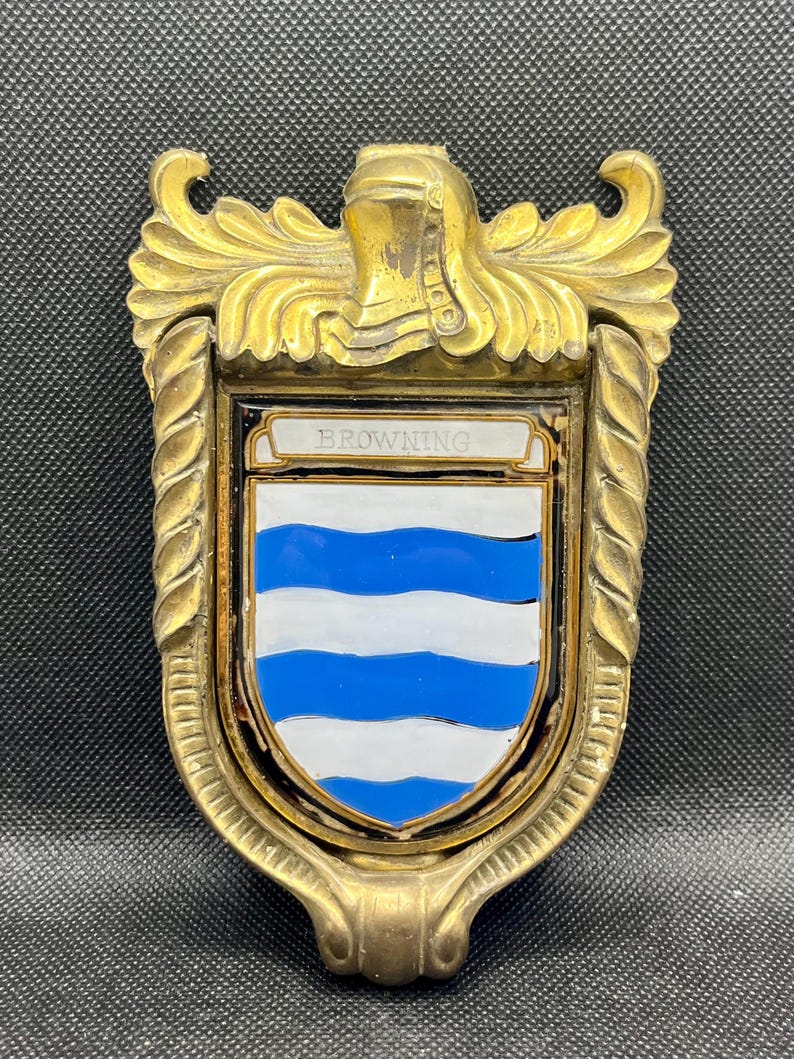 K&ouml;nnte beinhalten: Ein T&uuml;rklopfer aus Messing mit einem schildf&ouml;rmigen Emblem, das ein wei&szlig;-blaues Wellenmuster aufweist. Im oberen Teil des Schildes steht der Schriftzug BROWNING auf wei&szlig;em Grund. Der Rahmen ist mit einem Helmzier und gedrehten S&auml;ulen verziert.