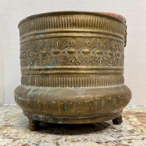 May include: A large, antique brass planter with an ornate design. The planter has a rounded base and three feet. The planter is made of brass and has a patina finish.