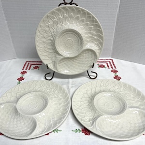 May include: Three off-white ceramic serving plates, each featuring a central circular bowl surrounded by a textured petal pattern and a separate kidney-shaped compartment. One plate is displayed upright on a black metal stand.