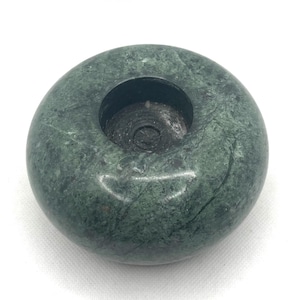 May include: A circular, green marble candle holder with a central circular recess. The stone displays a mottled pattern of dark and light green shades. This candle holder is a decorative item for home decor.