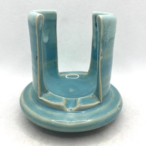 May include: A light blue ceramic sponge holder with a glossy finish and a U-shaped opening. The piece features a circular base and vertical sides with a lighter, mottled glaze near the top edges, creating a smooth, rounded, and functional form.