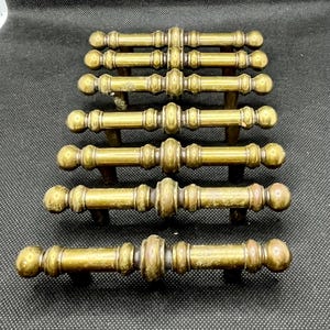 May include: A collection of seven antique brass drawer pulls. Each pull features a central decorative element flanked by smaller, detailed sections and rounded end knobs. The brass has a warm, aged patina, suggesting vintage craftsmanship. These pulls are designed for furniture restoration.