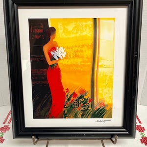 May include: Framed art print featuring a woman in a red dress holding white flowers. The artwork has a vibrant color palette of red, yellow, and green. The black frame is displayed on a decorative metal stand.