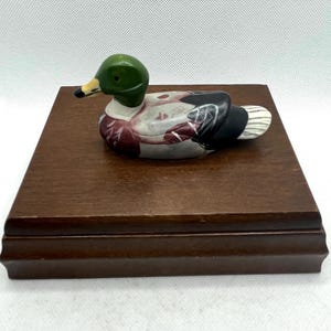 May include: A decorative duck figurine with a green head, white and maroon body, and black wings rests on a dark brown wooden base. The duck has a yellow beak and is positioned horizontally.