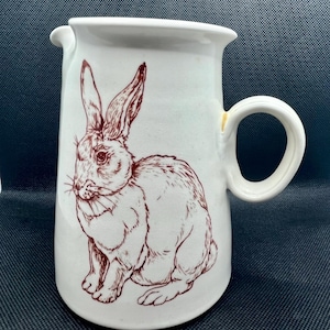 May include: White ceramic pitcher with a brown illustration of a rabbit. The pitcher has a rounded handle and a spout for pouring. The rabbit is depicted in a detailed, sketched style.
