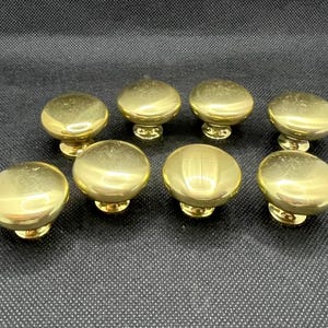 May include: Eight shiny, gold-colored cabinet knobs arranged on a dark surface. Each knob has a rounded top and a decorative base. The knobs are made of metal and reflect light.
