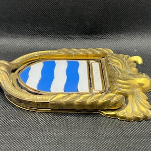 K&ouml;nnte beinhalten: Ein goldfarbener T&uuml;rklopfer aus Metall mit einem verzierten, seilartig strukturierten Rahmen und einem zentralen Wappenschild. Das Schild zeigt abwechselnd horizontale, wellenf&ouml;rmige blaue und wei&szlig;e Streifen sowie eine kleine rechteckige Plakette mit der Aufschrift BROWNING.
