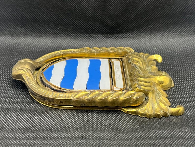K&ouml;nnte beinhalten: Ein goldfarbener T&uuml;rklopfer aus Metall mit einem verzierten, seilartig strukturierten Rahmen und einem zentralen Wappenschild. Das Schild zeigt abwechselnd horizontale, wellenf&ouml;rmige blaue und wei&szlig;e Streifen sowie eine kleine rechteckige Plakette mit der Aufschrift BROWNING.
