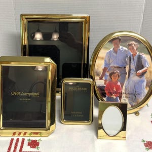 May include: A collection of five polished gold-tone brass picture frames in various shapes and sizes. The set includes rectangular, oval, and small portrait frames, some featuring the text Carr International Solid Brass and The Solid Brass Collection.