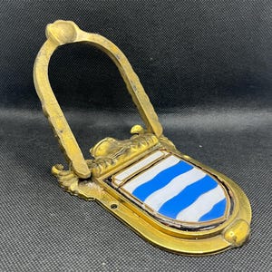 K&ouml;nnte beinhalten: Ein T&uuml;rklopfer aus Messing mit einem bogenf&ouml;rmigen Griff und einem schildf&ouml;rmigen Emblem. Der Schild zeigt abwechselnd blaue und wei&szlig;e horizontale Wellenstreifen mit dem Wort BROWNING auf einem kleinen wei&szlig;en Banner am oberen Rand.