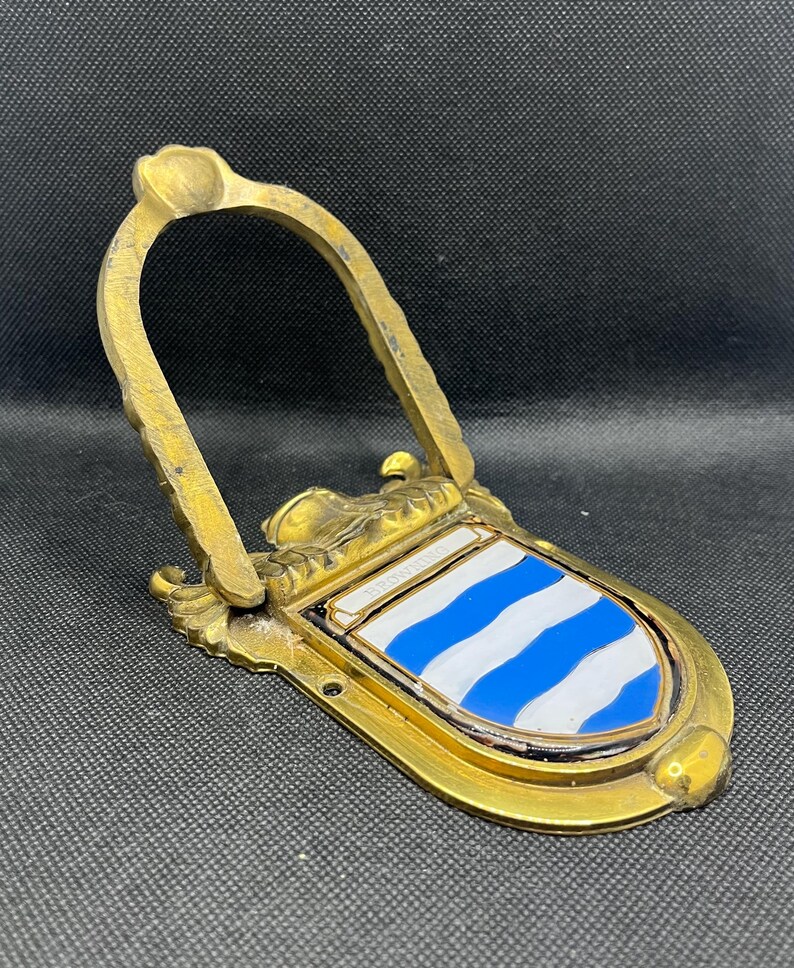 K&ouml;nnte beinhalten: Ein T&uuml;rklopfer aus Messing mit einem bogenf&ouml;rmigen Griff und einem schildf&ouml;rmigen Emblem. Der Schild zeigt abwechselnd blaue und wei&szlig;e horizontale Wellenstreifen mit dem Wort BROWNING auf einem kleinen wei&szlig;en Banner am oberen Rand.