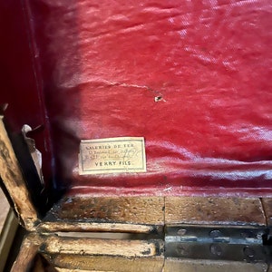 May include: Close-up of a red fabric-covered wooden box with a label that reads "Galeries De Fer, 6 Boulevard St. Martin, 35 rue de Charenton, VERRY FILS."