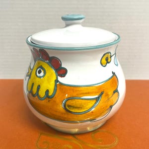 May include: A white ceramic sugar bowl with a lid, featuring a hand-painted yellow and orange chicken design with a red comb. The bowl has a light blue rim and a matching light blue knob on the lid, with a small yellow flower accent.