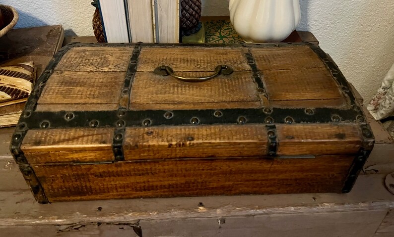 May include: A wooden chest with a brass handle and black metal accents. The chest has a distressed finish and is made of wood.