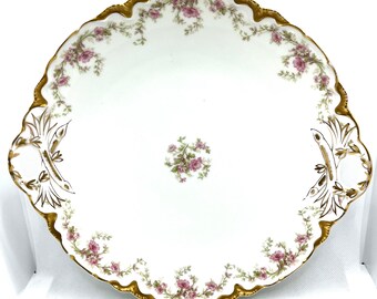 May include: A round white porcelain serving platter with a scalloped edge trimmed in gold. The plate features a central cluster of pink flowers and green leaves, with matching floral garlands along the rim and gold accents on the handles.