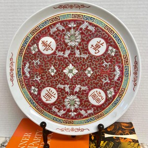May include: A white ceramic plate with a red and gold border. The centre of the plate features a red background with a white floral pattern and four Chinese characters. The characters are "福", "祿", "壽", and "喜", which translate to "happiness", "prosperity", "longevity", and "joy".