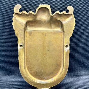 K&ouml;nnte beinhalten: Eine an der Wand montierbare Messingplakette mit einer schildf&ouml;rmigen Mitte und einer glatten, vertieften Oberfl&auml;che. Der &auml;u&szlig;ere Rand zeigt verzierte, symmetrische Schn&ouml;rkel und Dekorationen. Zwei kleine kreisf&ouml;rmige L&ouml;cher befinden sich an den Seiten.