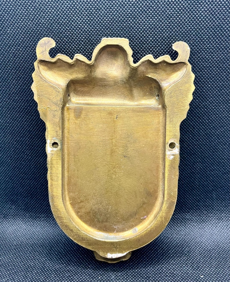 K&ouml;nnte beinhalten: Eine an der Wand montierbare Messingplakette mit einer schildf&ouml;rmigen Mitte und einer glatten, vertieften Oberfl&auml;che. Der &auml;u&szlig;ere Rand zeigt verzierte, symmetrische Schn&ouml;rkel und Dekorationen. Zwei kleine kreisf&ouml;rmige L&ouml;cher befinden sich an den Seiten.