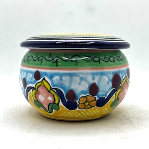 May include: A round, hand-painted ceramic jar with a lid. The jar features a vibrant design with yellow, blue, green, and pink floral patterns. The lid is dark blue with a white stripe. The jar is likely used for storage.