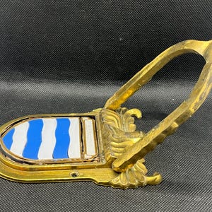 K&ouml;nnte beinhalten: Ein goldfarbener T&uuml;rklopfer aus Metall mit einem bogenf&ouml;rmigen Griff und einer schildf&ouml;rmigen Platte. Die Platte zeigt ein blau-wei&szlig;es Wellenstreifenmuster und enth&auml;lt einen kleinen rechteckigen Bereich mit der Gravur BROWNING.