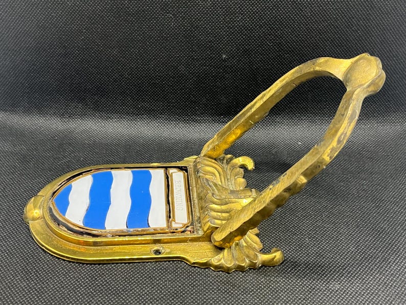 K&ouml;nnte beinhalten: Ein goldfarbener T&uuml;rklopfer aus Metall mit einem bogenf&ouml;rmigen Griff und einer schildf&ouml;rmigen Platte. Die Platte zeigt ein blau-wei&szlig;es Wellenstreifenmuster und enth&auml;lt einen kleinen rechteckigen Bereich mit der Gravur BROWNING.