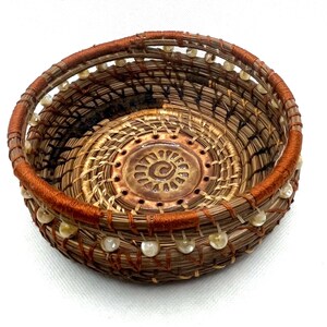 May include: A handcrafted, round basket with a spiral design in the center. The basket is made of natural fibers in shades of brown and tan, with a decorative orange rim and small, clear beads around the top edge.