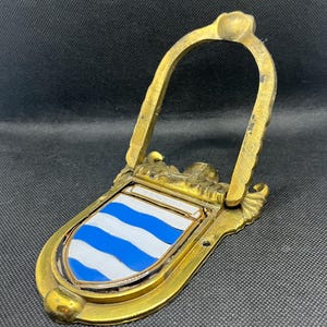 K&ouml;nnte beinhalten: Ein T&uuml;rklopfer aus Messing mit einem schildf&ouml;rmigen Emblem, das horizontale blaue und wei&szlig;e Wellenstreifen aufweist. Das Wort BROWNING ist auf einer kleinen rechteckigen Platte &uuml;ber dem Schild eingraviert. Mit klappbarem, bogenf&ouml;rmigem Griff.