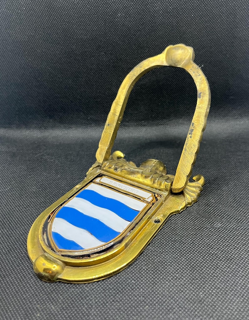K&ouml;nnte beinhalten: Ein T&uuml;rklopfer aus Messing mit einem schildf&ouml;rmigen Emblem, das horizontale blaue und wei&szlig;e Wellenstreifen aufweist. Das Wort BROWNING ist auf einer kleinen rechteckigen Platte &uuml;ber dem Schild eingraviert. Mit klappbarem, bogenf&ouml;rmigem Griff.
