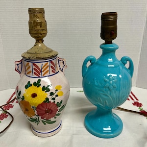 May include: Two ceramic table lamps. One is a light pink vase shape with a floral design in yellow, red, and orange. The other is a turquoise blue vase shape with a floral relief design. Both have brass lamp sockets.