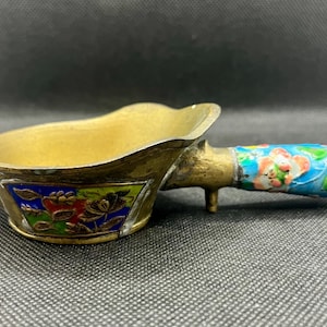 May include: A small brass scoop with a handle featuring a blue, pink, and green floral enamel pattern. The bowl of the scoop has a triangular inset panel decorated with a colorful floral design in red, blue, and green enamel on a brass base.