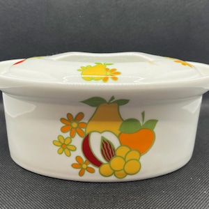 May include: White ceramic casserole dish with a lid, decorated with colorful fruit illustrations including pears, peaches, and oranges. The dish has a rounded shape and a scalloped edge on the lid. The fruit designs are in shades of yellow, orange, red, and green.
