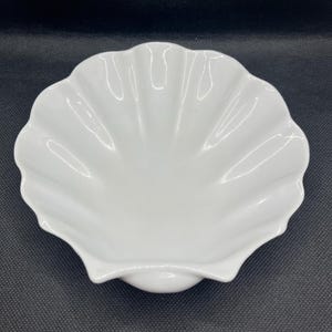 May include: A white ceramic bowl shaped like a scallop shell. The bowl features a smooth, glossy finish with deep, radiating ridges that extend from the center to the scalloped edges, creating a distinct shell-like appearance.