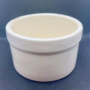 May include: A small, off-white ceramic pot with a rounded body and a thick, slightly textured rim. The interior is a lighter cream colour. The pot is designed for planting small plants or as a decorative item.