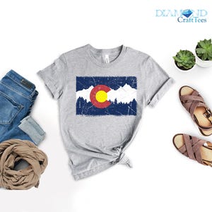 May include: A grey t-shirt displaying the Colorado state flag. The flag design features a red circle with a yellow centre, a blue rectangle, and white mountain and tree silhouettes. The shirt is styled with blue jeans, a scarf, and sandals.
