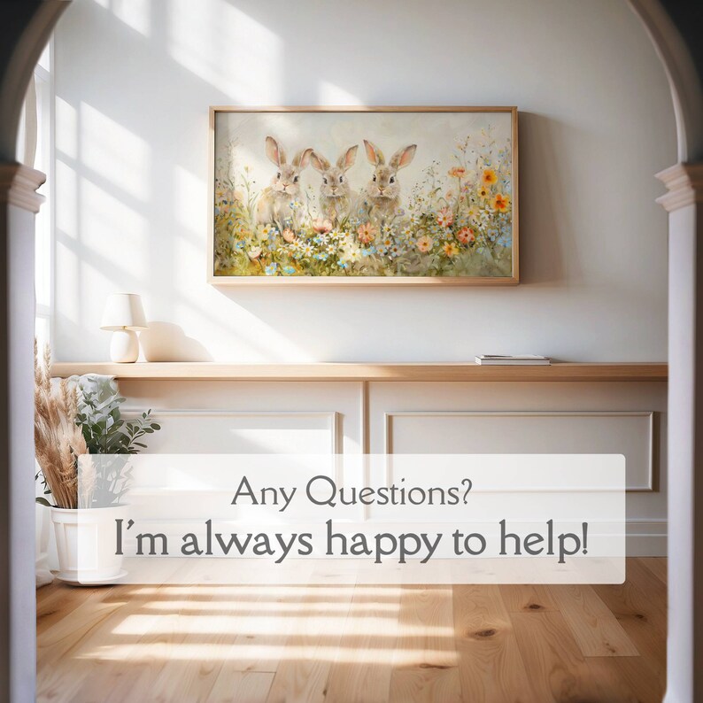 May include: A framed painting of three brown rabbits sitting in a field of colorful flowers. The painting is hanging on a white wall in a hallway with a wooden floor.