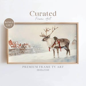 May include: A watercolor painting of a reindeer with a red and green garland around its neck standing in a snowy village scene. The reindeer is looking to the left of the image. The village is in the background and is made up of small houses with red roofs. The painting is framed in a light wood frame.