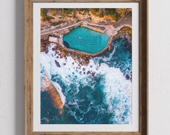 以下が含まれることがあります： 岩の崖と砕ける波に囲まれたターコイズブルーの海水のプールを上空から見た写真です。プールには泳いでいる人たちがいます。