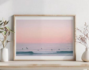 以下が含まれることがあります： 穏やかな海と小さな波、そして柔らかなピンクとピーチ色の夕焼けが描かれた横長の風景画の額縁入りプリント。海面にはサーファーの暗いシルエットがいくつか見えます。フレームは明るい木製です。